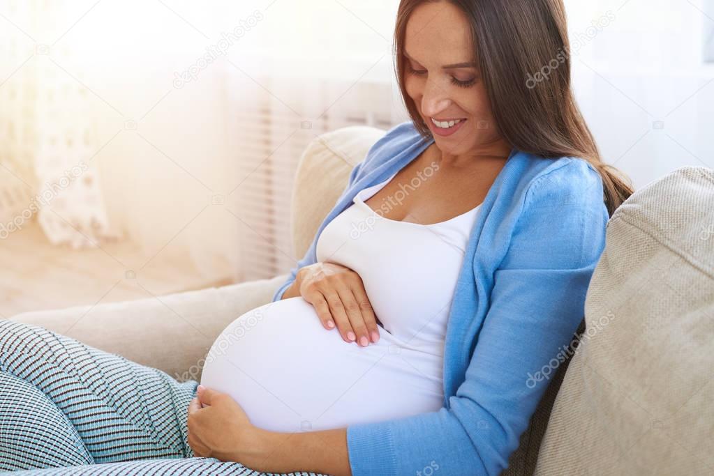 Pregnant woman holding her belly with arms on sofa 