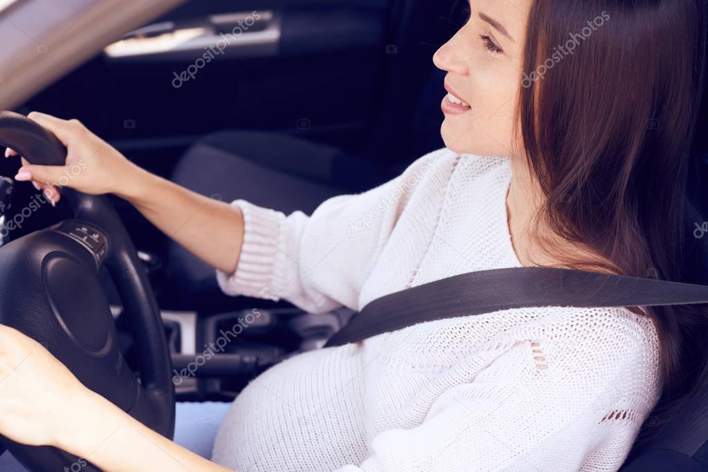 Happy pregnant female driver steering car with safety belt