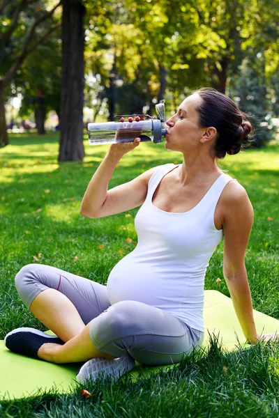 Вагітна жінка п'є воду на йога мат в парку — стокове фото
