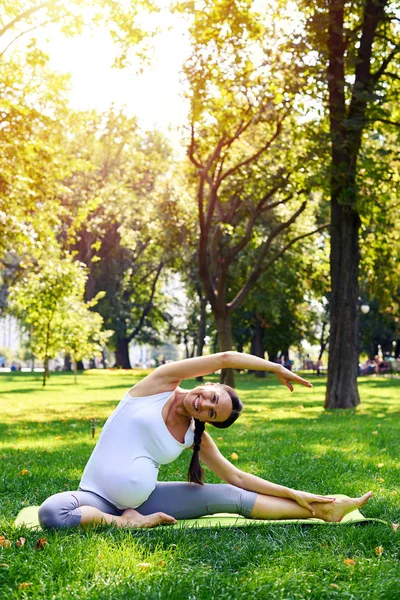 Sorrindo mulher grávida esticando no tapete de ioga no parque — Fotografia de Stock