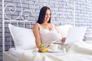 Dizüstü bilgisayar kullanarak ve macaroons çay ile yatakta yeme hamile kadın