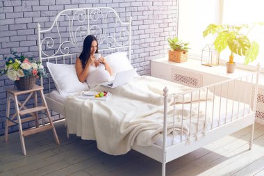 Dizüstü bilgisayar kullanarak ve yatakta kahvaltı hamile kadın