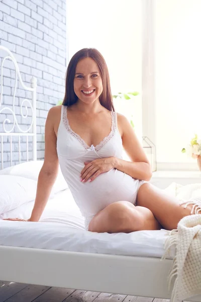 Беременная женщина прикасается к животу, сидя на кровати — стоковое фото