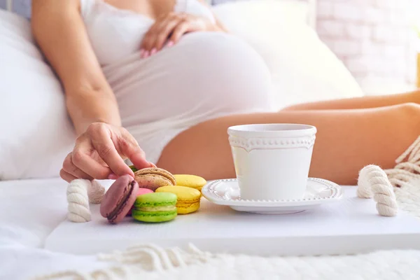 Hand einer schwangeren Frau, die eine Makrone auf dem Bett hält — Stockfoto