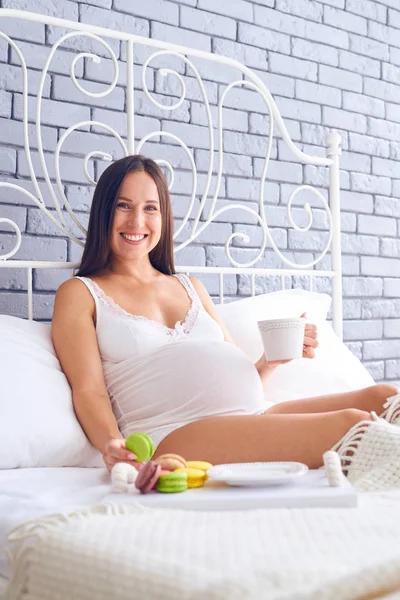 Lächelnde Schwangere trinkt morgens eine Tasse Tee mit Makronen — Stockfoto