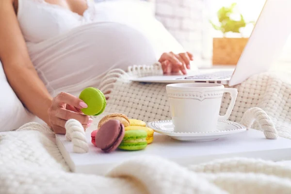 Hand av gravid kvinna som håller macaroon medan du använder laptop i vara — Stockfoto