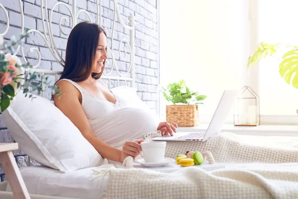 Femme enceinte avec ordinateur portable et plateau avec petit déjeuner assis dans être — Photo