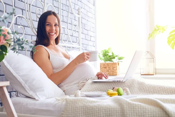 Mujer embarazada sonriente desayunando en la cama con computadora portátil — Foto de Stock