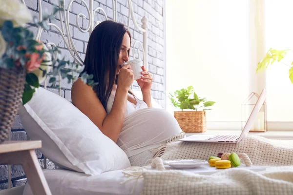Беременная женщина в пижаме пьет чай и пользуется ноутбуком на кровати — стоковое фото