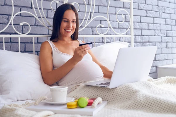 Mujer embarazada sonriente sentada en la cama con portátil y coche de crédito —  Fotos de Stock