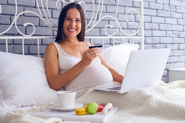 Hamile kadın yatakta laptop ve kredi kartı ile oturuyordu — Stok fotoğraf