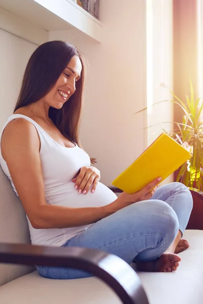 Έγκυος μελαχρινή γυναίκα χαλαρώνοντας στον καναπέ του, διαβάζοντας το βιβλίο — Φωτογραφία Αρχείου