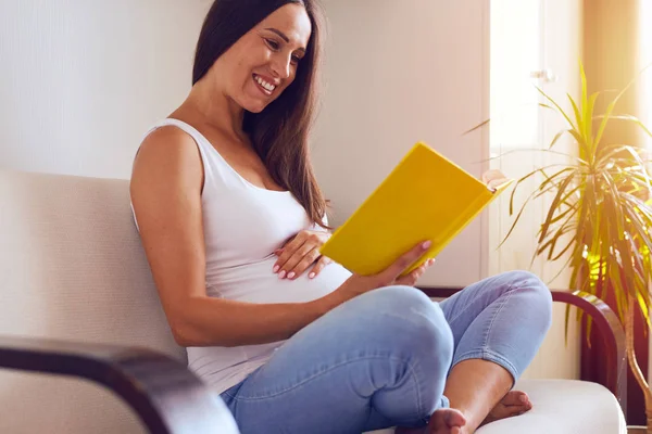 Happy pregnant woman reading book on sofa — Stock Photo, Image