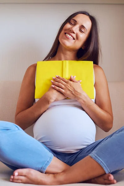 Ευχαριστημένος έγκυος γυναίκα, κρατώντας το βιβλίο ενώ κάθονται στον καναπέ — Φωτογραφία Αρχείου