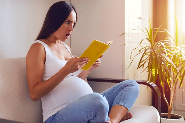 Mulher grávida surpreso ler livro enquanto sentado no sofá — Fotografia de Stock