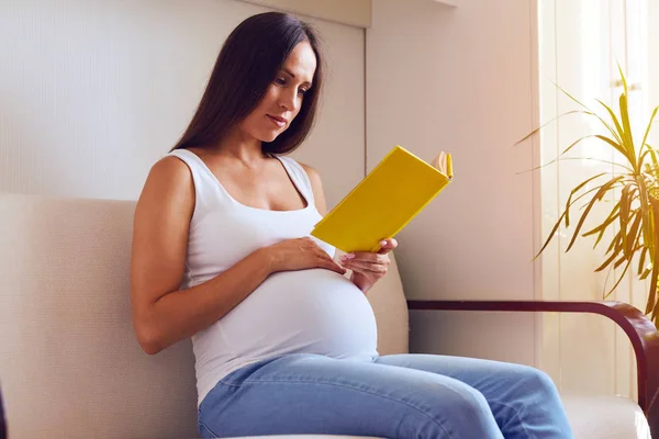 Mujer embarazada concentrada leyendo libro mientras está sentada en el sofá —  Fotos de Stock