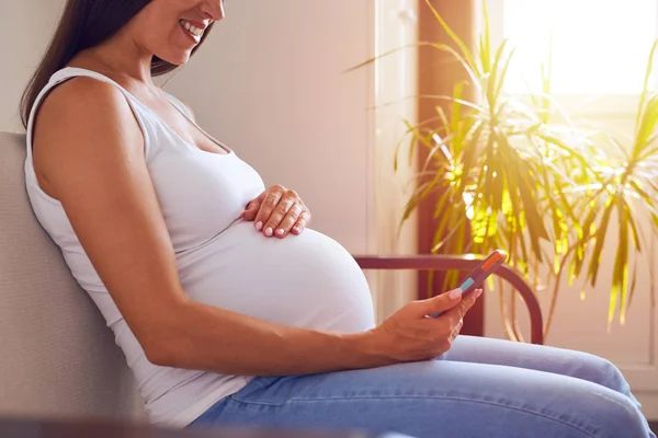 Mother-to-be sitting on sofa and using phone — Stock Photo, Image