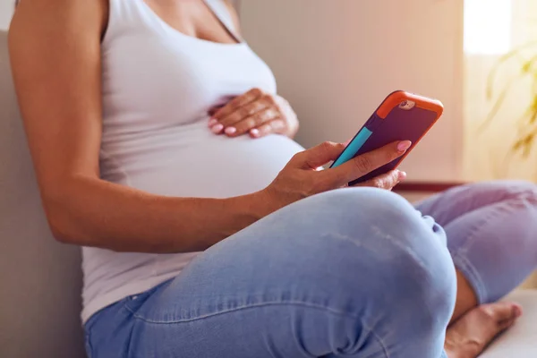 Mãos de fêmea tocando barriga grávida e usando telefone em casa — Fotografia de Stock