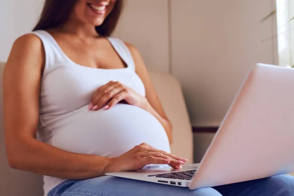 Беременная женщина с ноутбуком сидит дома на диване — стоковое фото