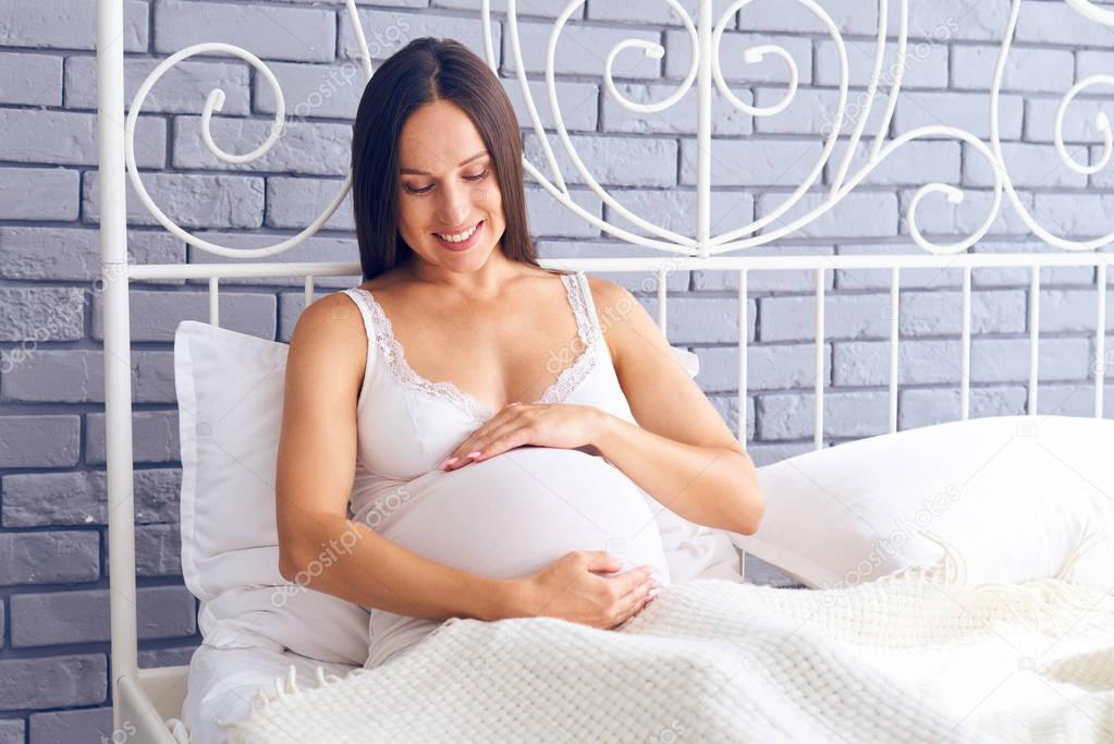Happy woman looking at pregnant belly on bed 