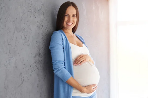 Sonriendo mujer embarazada tocando el vientre mientras mira a la cámara —  Fotos de Stock