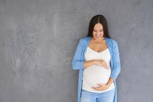 Mujer embarazada joven mirando su vientre contra el fondo gris —  Fotos de Stock