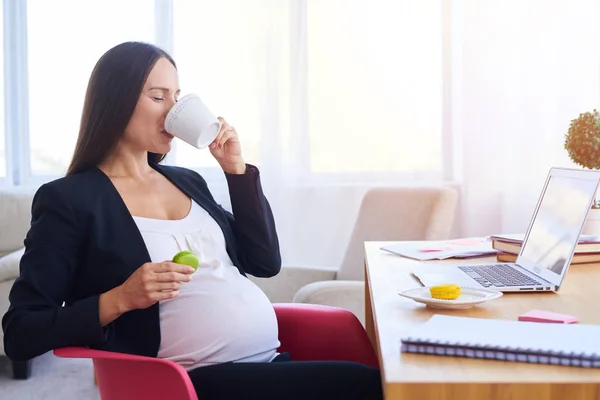 Pregnant businesswoman drinking tea and eating sweets at workpla — Stock Photo, Image