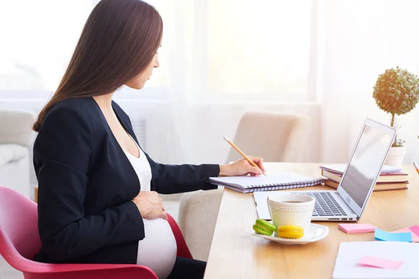 Pregnant woman holding hand on belly and making notes — Stock Photo, Image