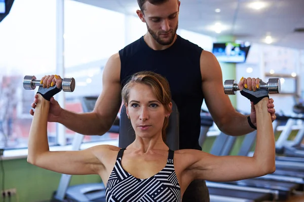 หนุ่มสาวที่มุ่งมั่นผู้หญิง flexing biceps กับครูฝึกส่วนตัวใน g — ภาพถ่ายสต็อก