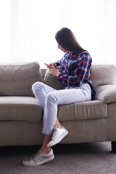 Ung flicka sitter på hem soffa och med mobiltelefon — Stockfoto