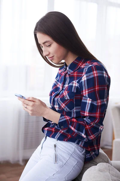 Chica joven sociable mensajes de texto en el teléfono inteligente en la sala de estar —  Fotos de Stock