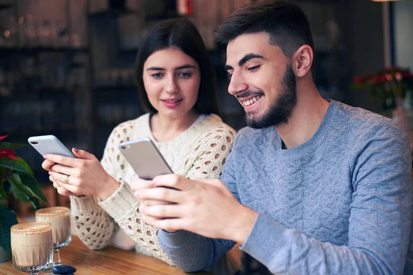 Alegre pareja de mensajes de texto a través de teléfonos mientras citas en la cafetería —  Fotos de Stock