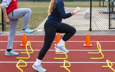 İki lise atletizm koşucusu koşuyor ve kırmızı bir pistte sarı mini engelleri aşıyor..