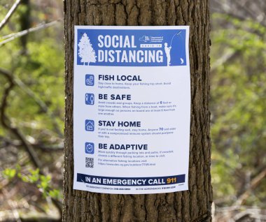 Babil, New York, ABD - 1 Nisan 2020: Parktaki bir ağaçta insanlara Sosyal Mesafe, yerel balıklar, güvende olun, evinizde kalın ve adapte olun diyen bir kağıt işareti var.
