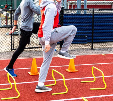 İki liseli çocuk atletizm antrenmanı sırasında kırmızı pistte sarı mini engelleri ve turuncu konileri aşıyor..