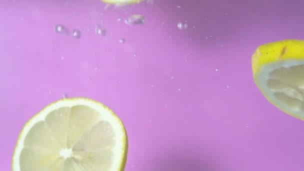Gros plan vidéo au ralenti de tranches de citron tombant et filant dans l'eau sur fond violet et turquoise. Il y a deux parties de clip .