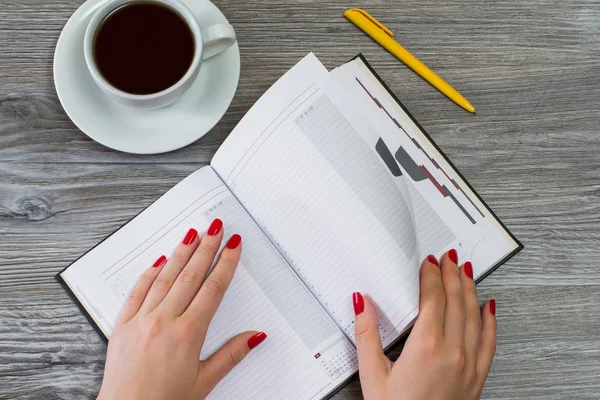 Woman's hands turning a page in a notebook. Cup of tea and a pen