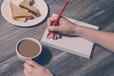 Bir insanın elini bir defterde yazılı. İş yerinde kır. Arka plan bir fincan sıcak kahve, lezzetli peynirli sandviç tutan bir el. Üstten Görünüm, vintage tarzı