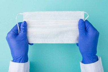 Doktorlar Coronavirus salgını sırasında güvende kalıyorlar. Yukarıdan bakıldığında, eldivenli doktorun elinde turkuaz arka planda izole edilmiş beyaz bir cerrahi maske olduğunu görüyoruz.