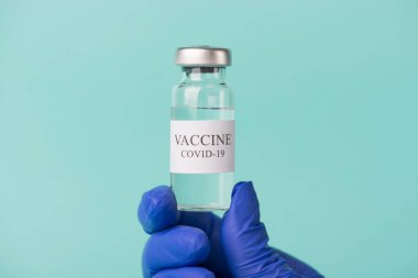 Yeni koronavirüs aşısı inceleme konsepti. Doktorun elinde mavi deniz mavisi arkaplanına izole edilmiş COVID-19 aşısı şişesi tutarken çekilmiş yakın çekim fotoğrafı.