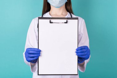 Mavi eldivenli, elinde boş bir pano olan, turkuaz deniz mavisi arkaplanından izole edilmiş, yakın planda çekilmiş bir kadın doktor fotoğrafı.