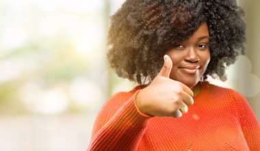Beautiful african woman smiling broadly showing thumbs up gesture to camera, expression of like and approval, outdoor clipart