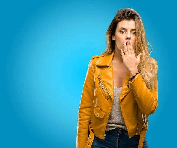 Beautiful young woman covers mouth in shock, looks shy, expressing silence and mistake concepts, scared, blue background