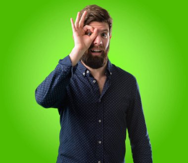 Parmakları Tamam jest olarak kameradan bakarak büyük sakallı genç hipster adam. Taklit dürbün, güzel gözleri ve gülümseme yeşil arka plan üzerinde