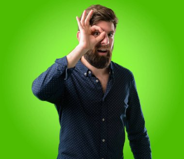 Parmakları Tamam jest olarak kameradan bakarak büyük sakallı genç hipster adam. Taklit dürbün, güzel gözleri ve gülümseme yeşil arka plan üzerinde