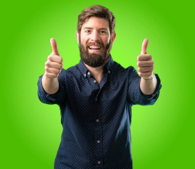 Genç hipster adam büyük sakal stand mutlu ve olumlu iyi jest yeşil arka plan üzerinde ifade kocaman bir gülümseme ile onaylama kadar baş parmakları ile