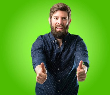 Genç hipster adam büyük sakal stand mutlu ve olumlu iyi jest yeşil arka plan üzerinde ifade kocaman bir gülümseme ile onaylama kadar baş parmakları ile