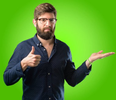 Genç hipster adam ile büyük sakal başparmak yukarıya bir şey boş elinde tutan ve yeşil arka plan üzerinde ok jest yapma