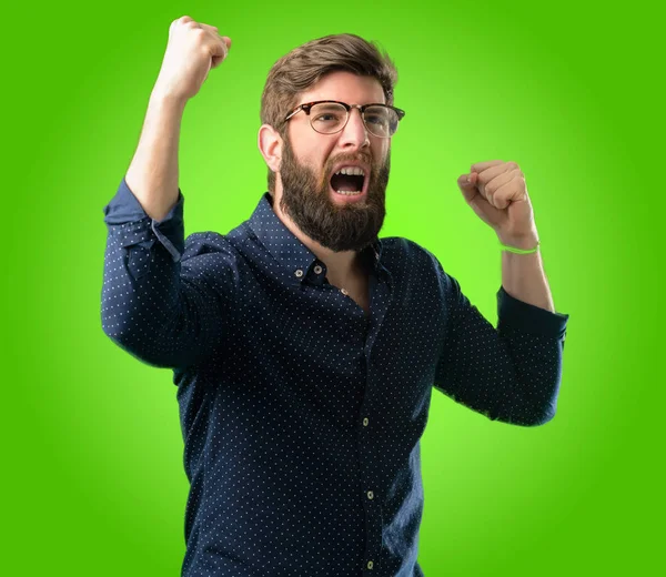 Young hipster man with big beard happy and excited celebrating victory expressing big success, power, energy and positive emotions. Celebrates new job joyful over green background