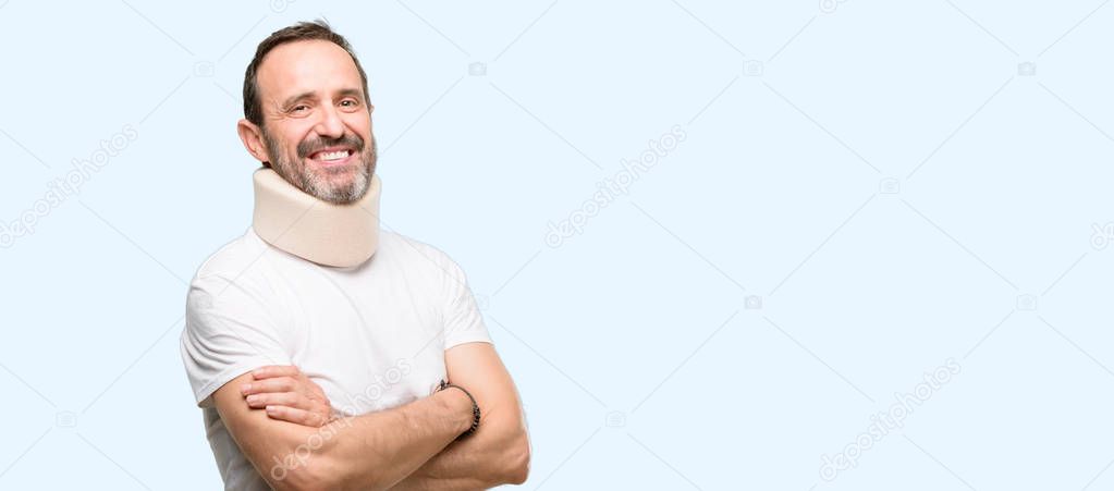 Injured senior man using a neck brace with crossed arms confident and happy with a big natural smile laughing isolated over blue background